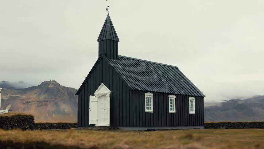 A solitary black church with a white door and trim stands in a barren landscape against a cloudy sky with a low stone wall surrounding it and distant mountains in the background