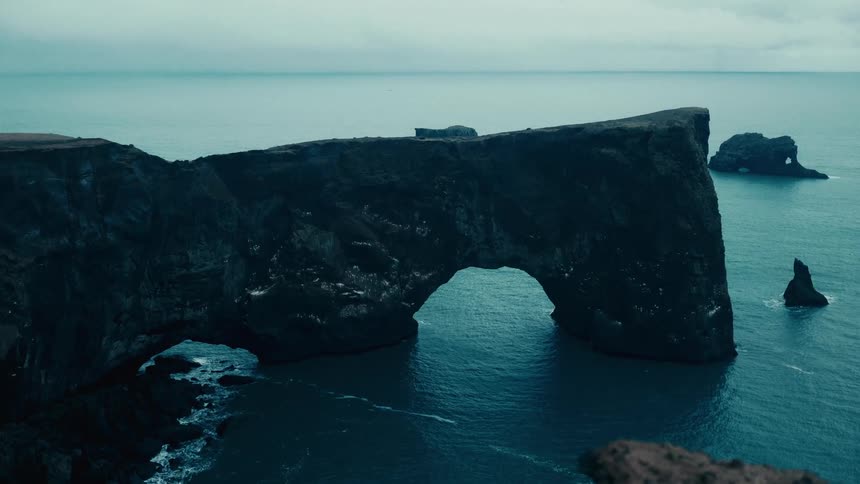 A large natural rock arch formation extends into the ocean with rugged cliffs and a calm sea in the background