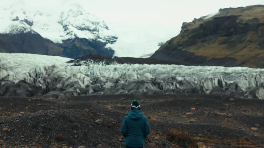 A person in a blue jacket and beanie stands on rocky terrain facing a vast glacier with snowcapped mountains in the background