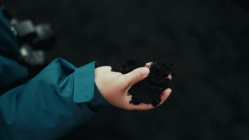 A hand in a blue sleeve is holding a handful of dark soil against a dark background