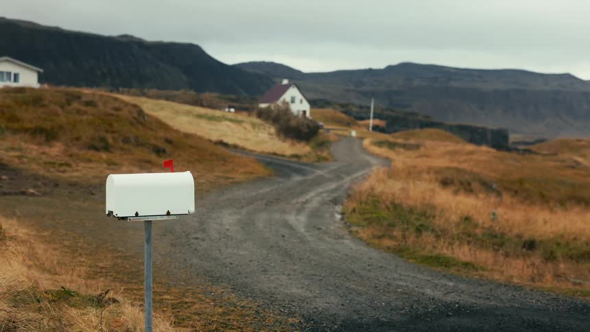 A white mailbox stands at the start of a winding gravel road leading to two houses nestled in a hilly brown and green landscape under a cloudy sky
