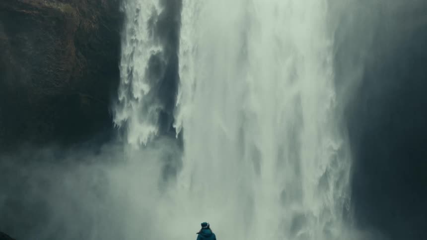 Person in a blue jacket enjoying the view of an imposing waterfall in front of him