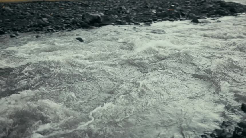 A fastflowing river with turbulent water runs over a rocky bed surrounded by dark wet stones and sparse vegetation in the background