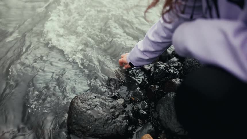 A woman wearing a jacket touches the surface water of a flowing river while standing on the rocky shore