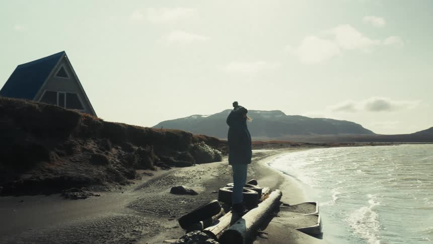 A person stands on a log near the shore of a beach with a small aframe house on a hill in the background and mountains visible in the distance