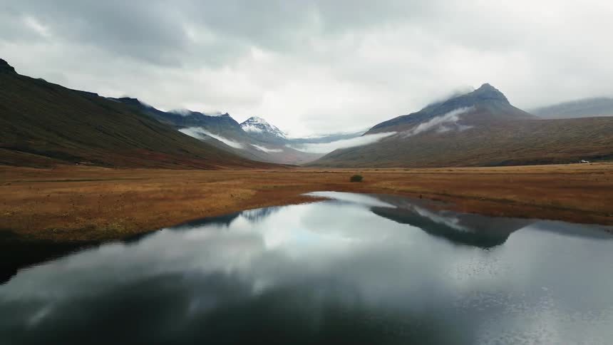 A still dark lake reflects the cloudy sky and surrounding brown and gray mountains