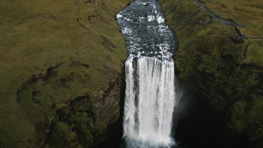 A dramatic aerial view of a powerful waterfall cascading down a cliff with turbulent water flowing through a narrow river flanked by lush green hills