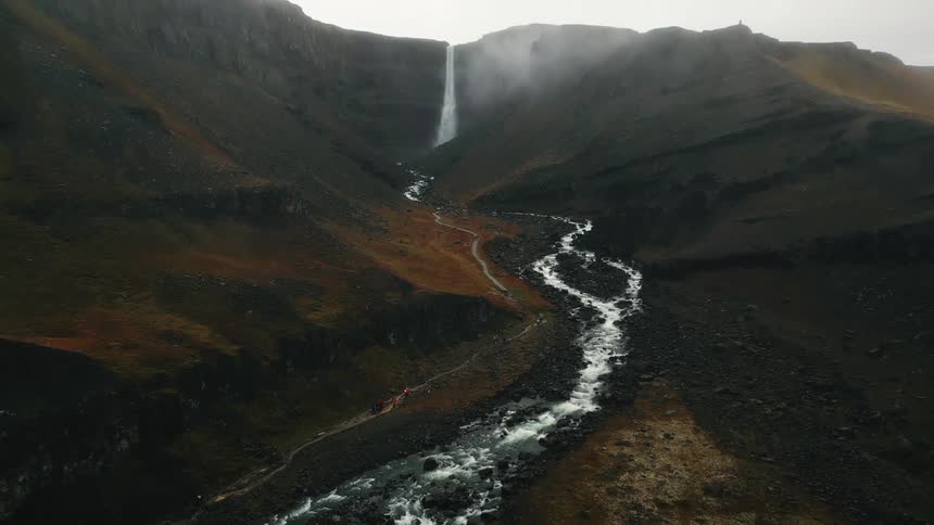 A tall thin waterfall plunges down a steep rocky cliff face bisecting a rugged mountain valley with a winding stream below