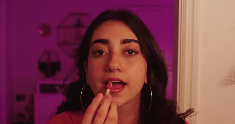 The face of a young woman who is looking into a mirror while applying lipstick to her lip and she is wearing hoop earrings