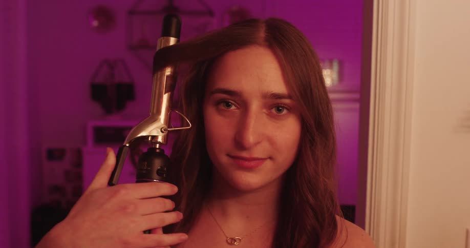 A girl with long hair is holding a curling iron styling her hair in a welllit room