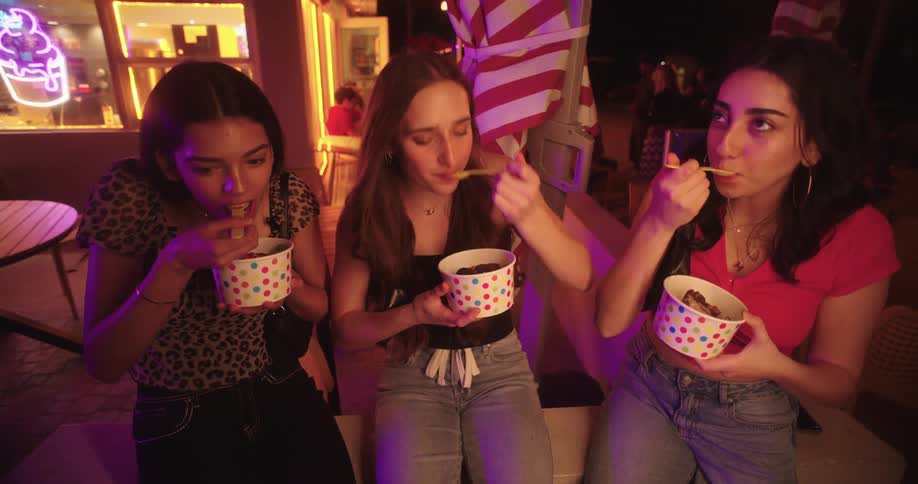 Three women are sitting next to each other and they are eating ice cream from white tubs with colorful dots