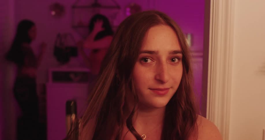 A teenage girl uses a curling iron to style her hair in a room while other teenage girls dance in the background