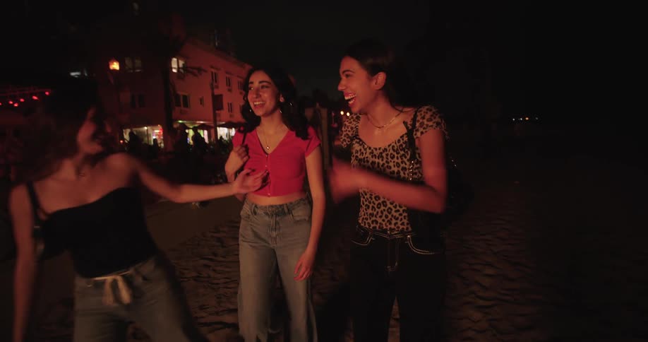 Three friends walking on the sand of the beach happy at night next to bars