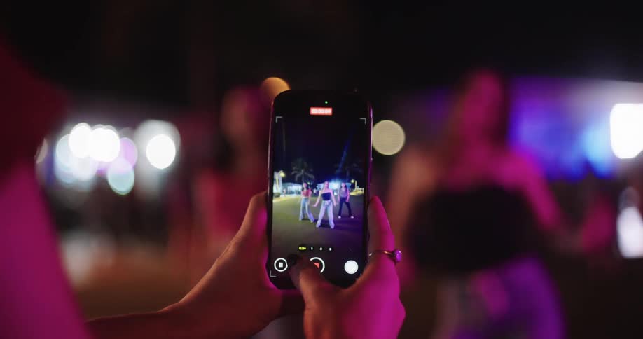 A person is holding a smartphone and taking a photo or video of a group of people dancing or performing under colorful lights at night