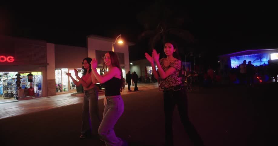 A teen is holding a smartphone and taking a video of a group of teenage girls dancing or performing under colorful lights at night