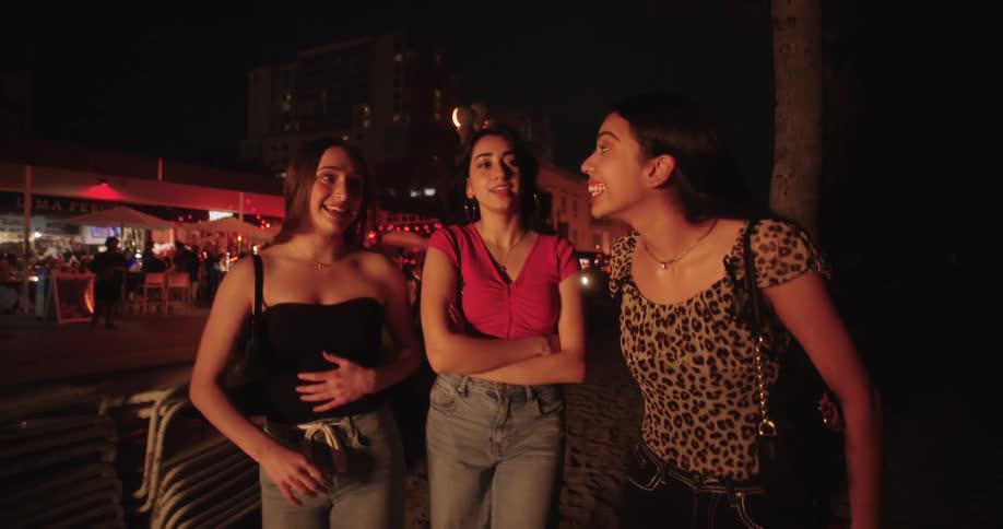 A group of three friends who are walking on a sand in a night and urban area and they are talking and smiling
