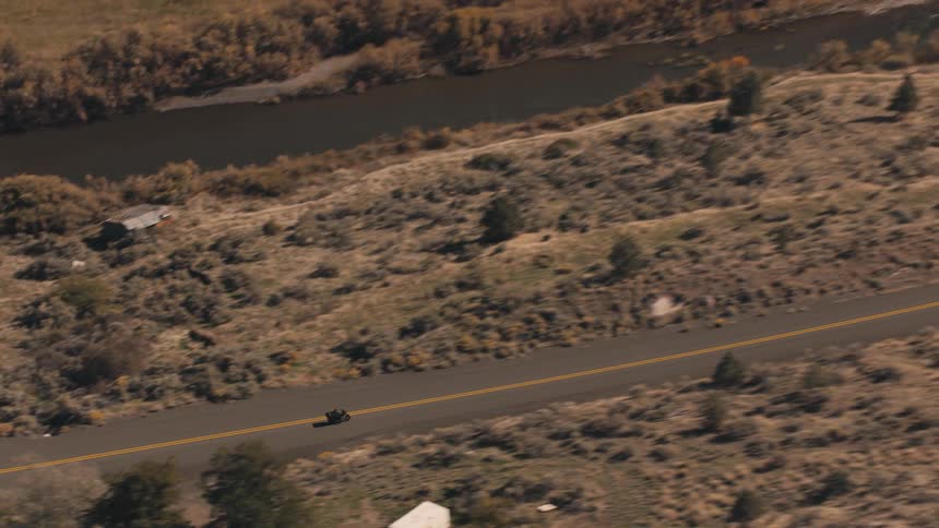 An aerial view zooms out showing a motorcycle driving on a rural road a white vehicle passes revealing a river and sparse buildings