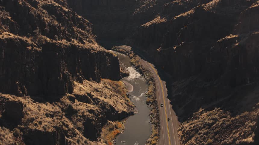 Vehicles travel along a highway paralleling a river passing between mountains on a sunny day