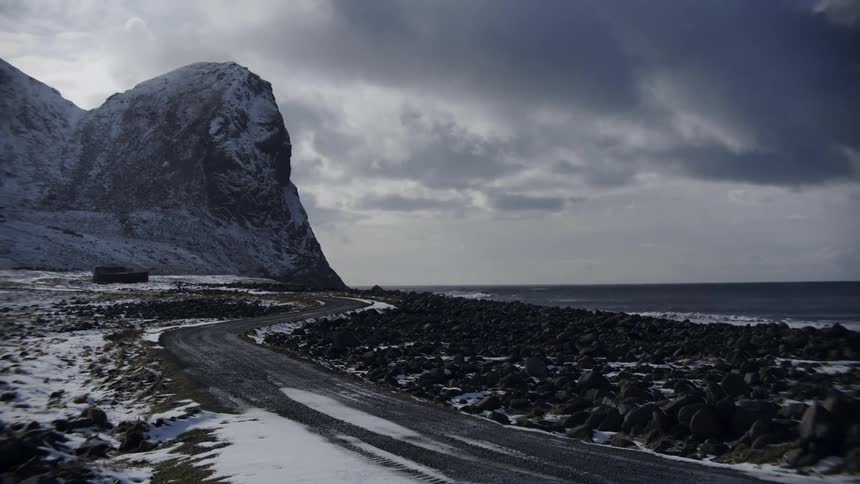 A winding road leads through a snowy landscape towards a rocky beach and a dramatic cloudy sky