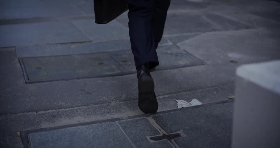 Legs of a person wearing dress shoes walking on a concrete paved street