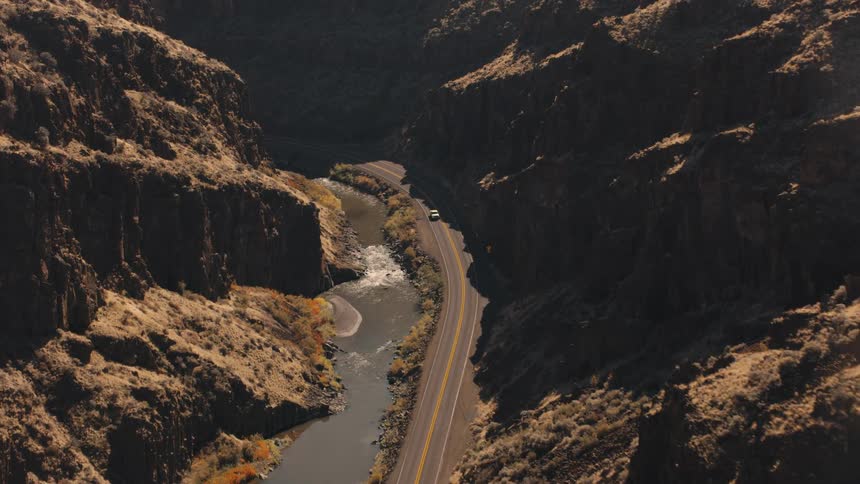An aerial view shows a white truck driving on a valley road alongside a flowing river nestled between steep cliffs