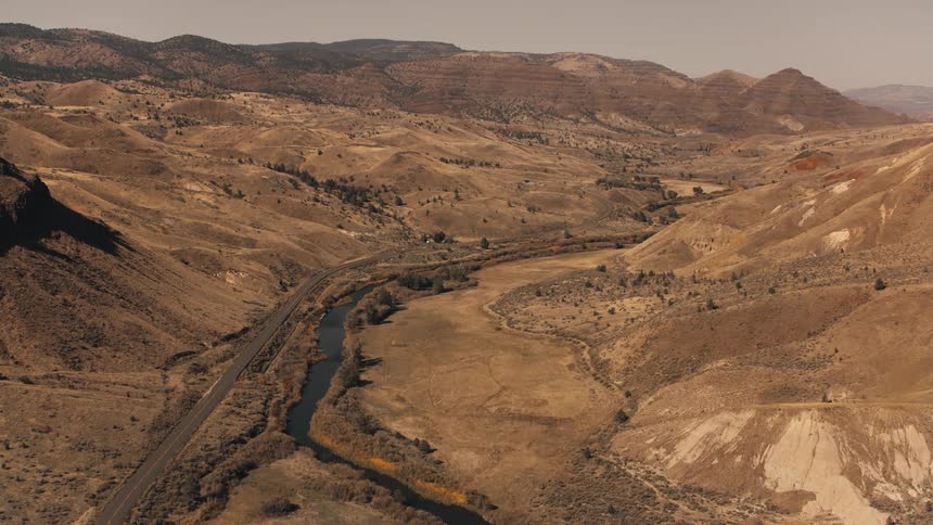 An expansive arid landscape with rolling hills a winding river sparse vegetation and a road running parallel to the river set against a backdrop of distant mountains