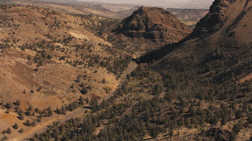 A winding road cuts through a vast arid landscape of rolling hills and sparse vegetation under a clear sky