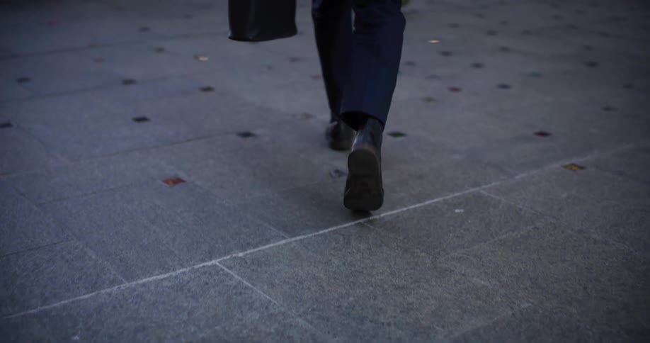 A person in dark pants and boots walks on a gray tiled surface with embedded colorful squares carrying a black bag