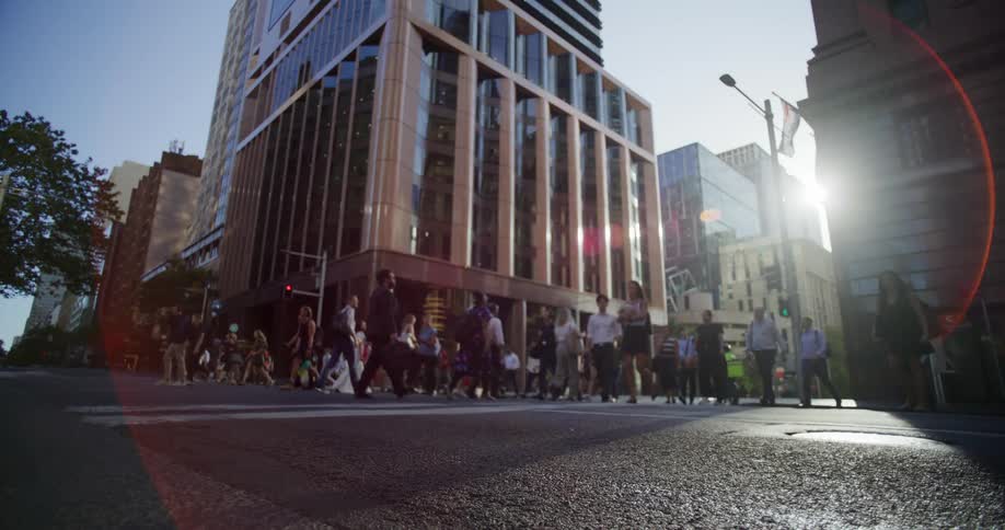 Pedastrians crossing a busy city street framed by tall buildings and a bright sunlit sky