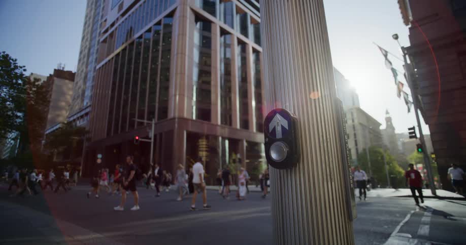 A pedestrian crossing button on a pole in a busy urban intersection with pedestrians crossing the street