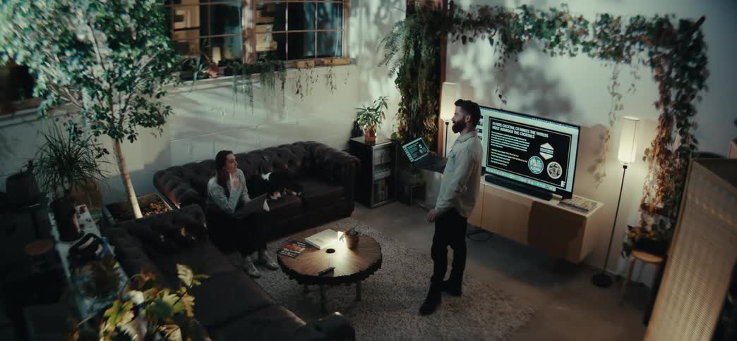 A cozy modern living space with lush indoor plants where a bearded man is giving a presentation using a laptop while a woman sits on a dark couch with a laptop and a black and white dog beside her