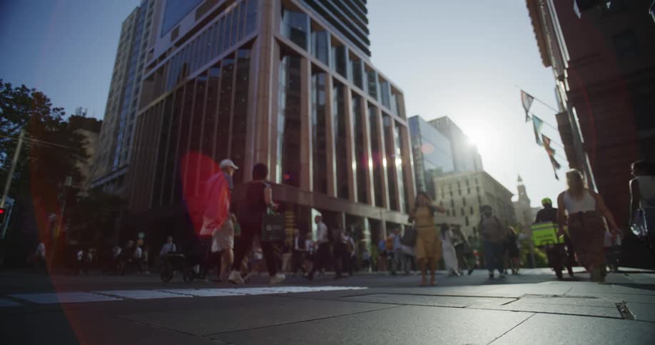 A busy urban street scene with people walking set against the backdrop of modern highrise buildings and the sun shining brightly in the sky