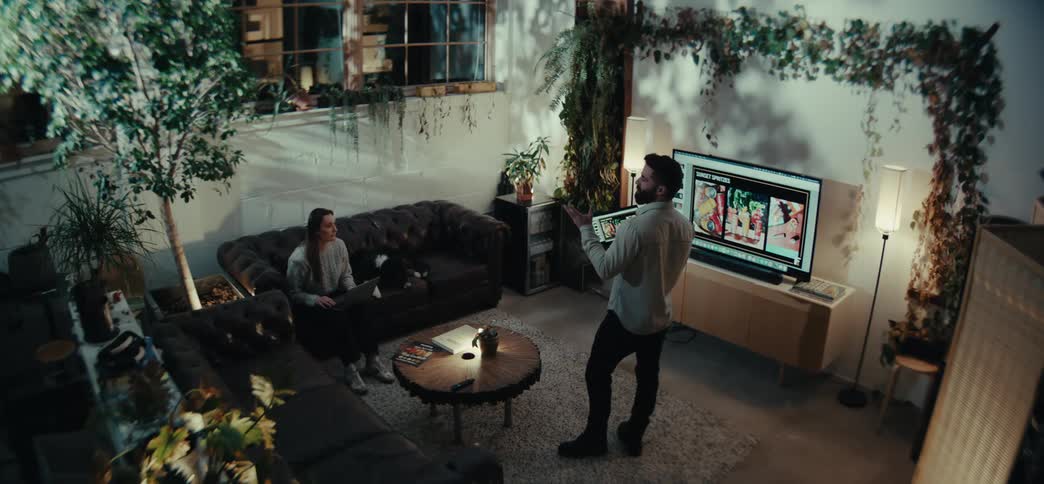 Man standing holding a laptop talking to a woman who is sitting in an armchair in front of him