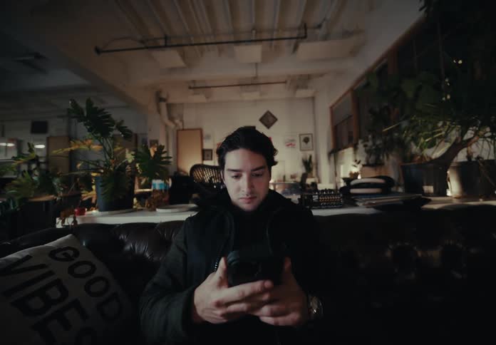 A person is sitting on a dark brown leather couch in a dimly lit room filled with various plants and decorations holding a smartphone with both hands