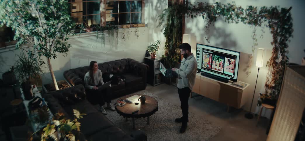 Man standing talking to a woman while she is sitting and he is holding a laptop