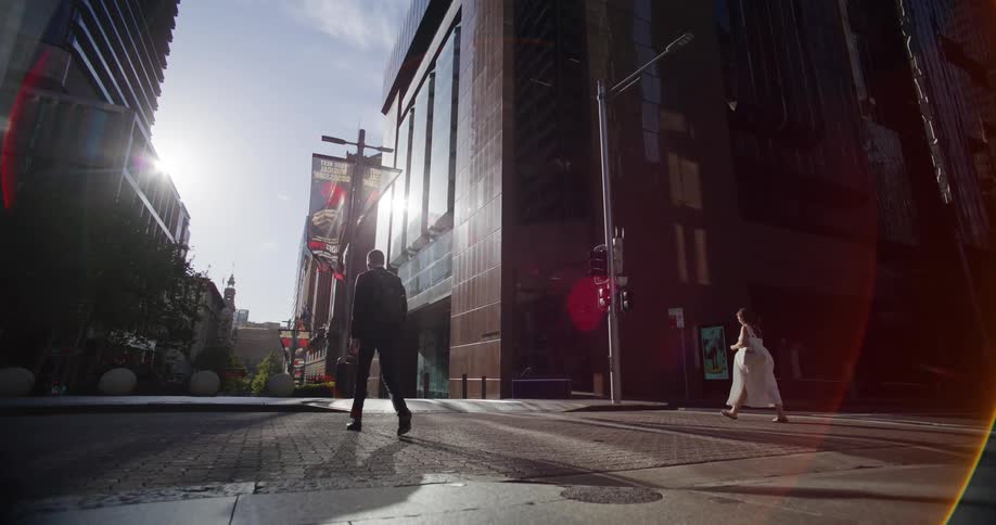 In a sun drenched cityscape with towering skyscrapers a man in a suit walks away from the camera down a cobblestone street while a woman in a flowing white dress walks along the sidewalk towards a traffic light