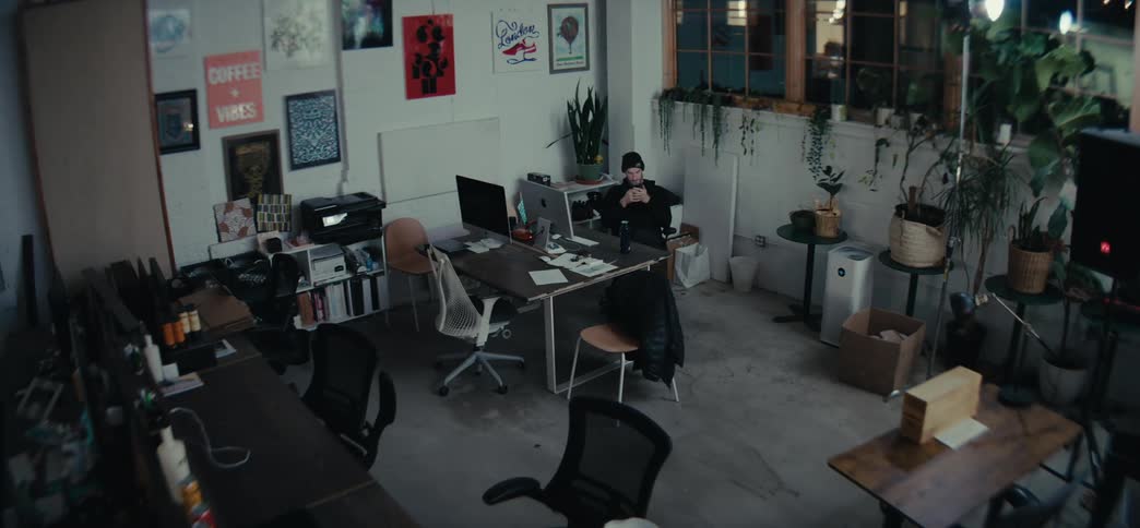 A man wearing a black hat and sweater sits at a desk in a cluttered industrialstyle office space illuminated by a warm lamp with various pieces of artwork and posters adorning the walls