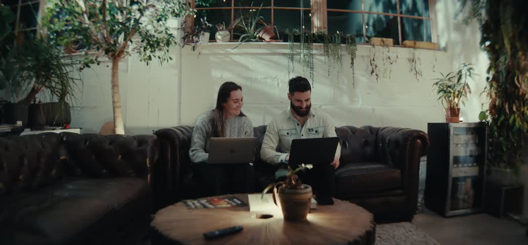 Man and woman sitting on a leather sofa using laptops in a welllit room with plants around