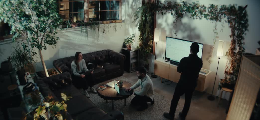 Three people in a relaxed work environment with plants around sitting and interacting with laptops and a television screen