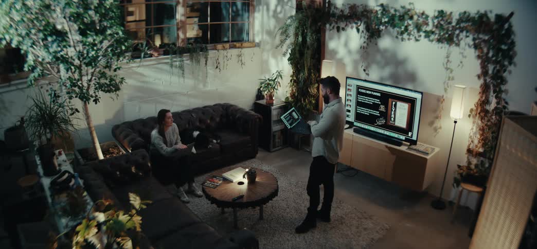 Man showing a laptop with graphics to a woman sitting on a sofa in a room with plants and a tv screen displaying information