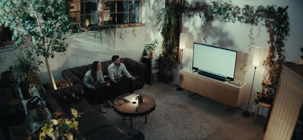 A modern living room with lush indoor plants where two people sit on a dark leather couch watching television