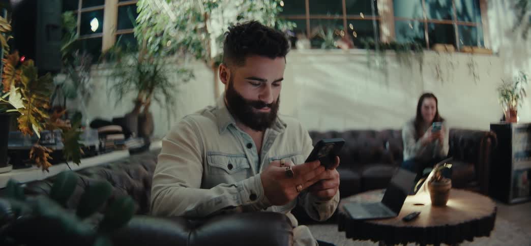 A bearded man in a denim jacket sitting on a leather couch focused on his phone while a woman in the background also holding a phone sits on another couch with a laptop open on a wooden table nearby in a cozy plant filled indoor setting