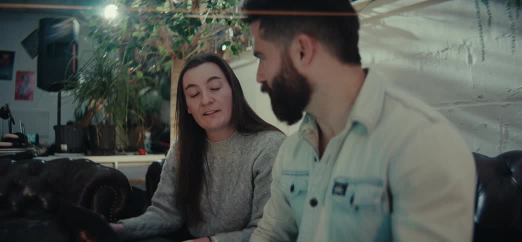 A man and a woman engaged in a conversation in a casual indoor setting with cozy furniture and plants in the background both have laptops on their laps with the woman looking attentively at the man while he gestures as he speaks