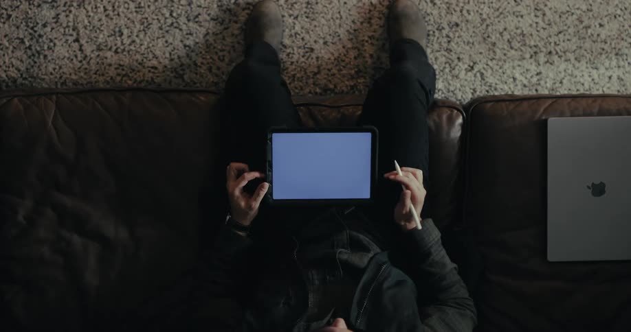 A man sits on a couch seen from an overhead perspective holding a tablet while a laptop rests beside them