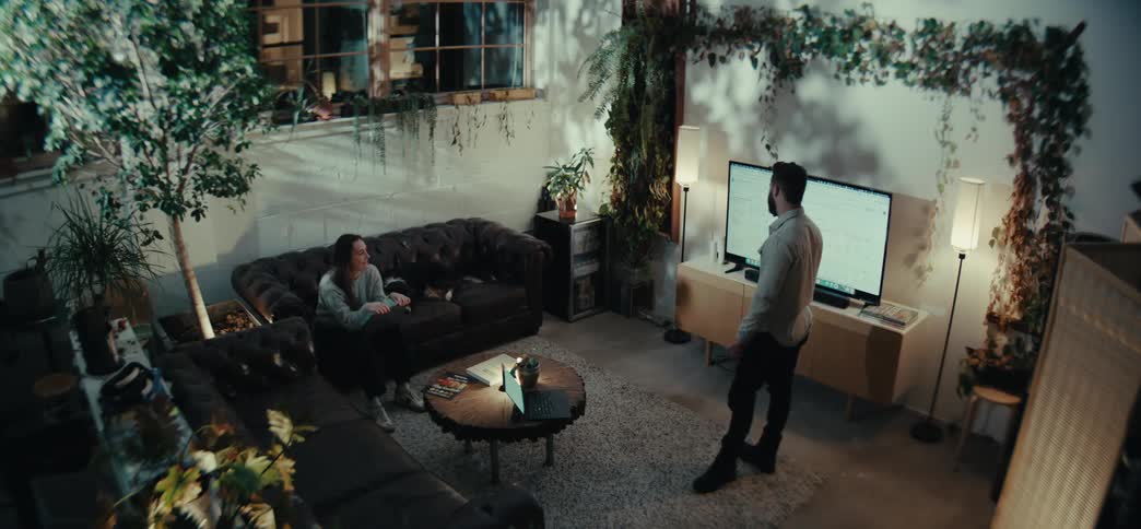 Man consults his laptop then gestures at a television screen while talking to a woman sitting in the living room at night