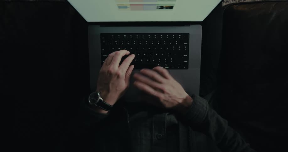 From above hands wearing a watch type on a laptop keyboard illuminated by the screens light against a dark background