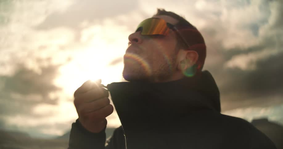 A man wearing sunglasses stands outdoors in front of a bright cloudy sky while wearing a dark jacket