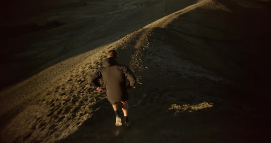 A man dressed in dark clothing runs along a rugged sandy ridge in a vast dramatic desert landscape with towering rock formations in the background