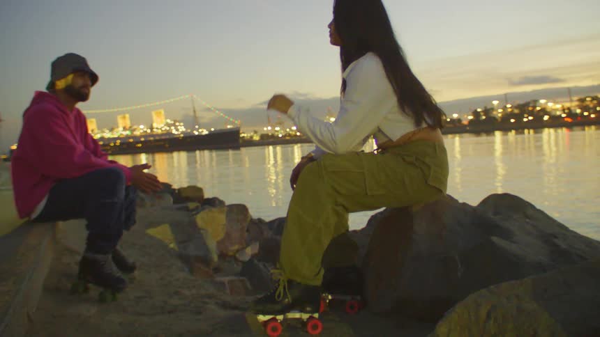A man and a woman are sitting on rocks that are in front of a body of water with an illuminated city in the background