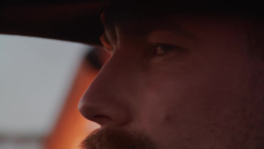 A mustached man wearing a cowboy hat stands near the burning flames his gaze fixed elsewhere